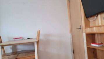 A room with a wooden desk, a chair, a television mounted on the wall, and a shelf with a red box.