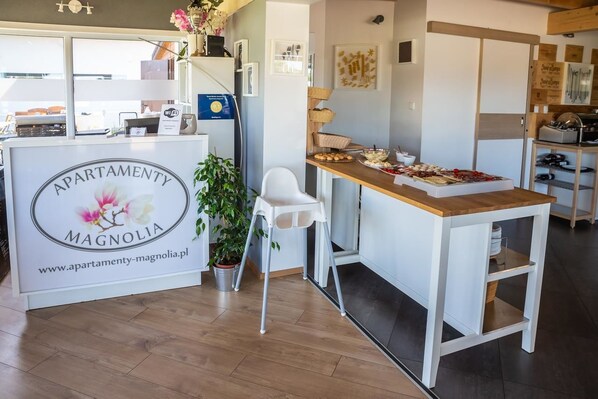 Zona de recepción con una mesa de madera que presenta aperitivos, una silla blanca y una planta en maceta.