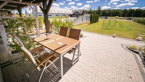 An outdoor patio with a wooden table and chairs, a pergola with climbing plants, and a view of a residential area with a lawn and a goalpost. - Apartamenty Magnolia (Wicko)