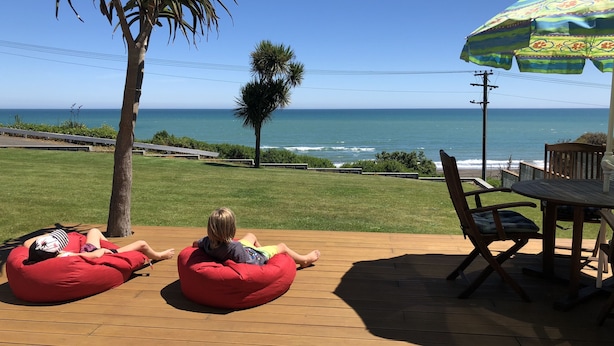 Une terrasse en bois avec deux fauteuils en forme de sac et une table avec des chaises, donnant sur une pelouse et l’océan.
