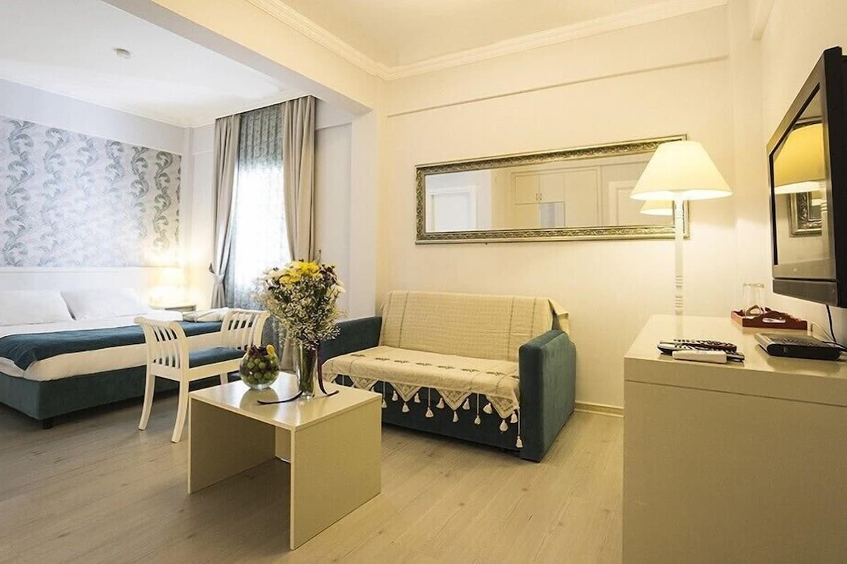 A hotel room with a bed, a sofa, a small table with a vase of flowers, a lamp, a mirror, and a TV.