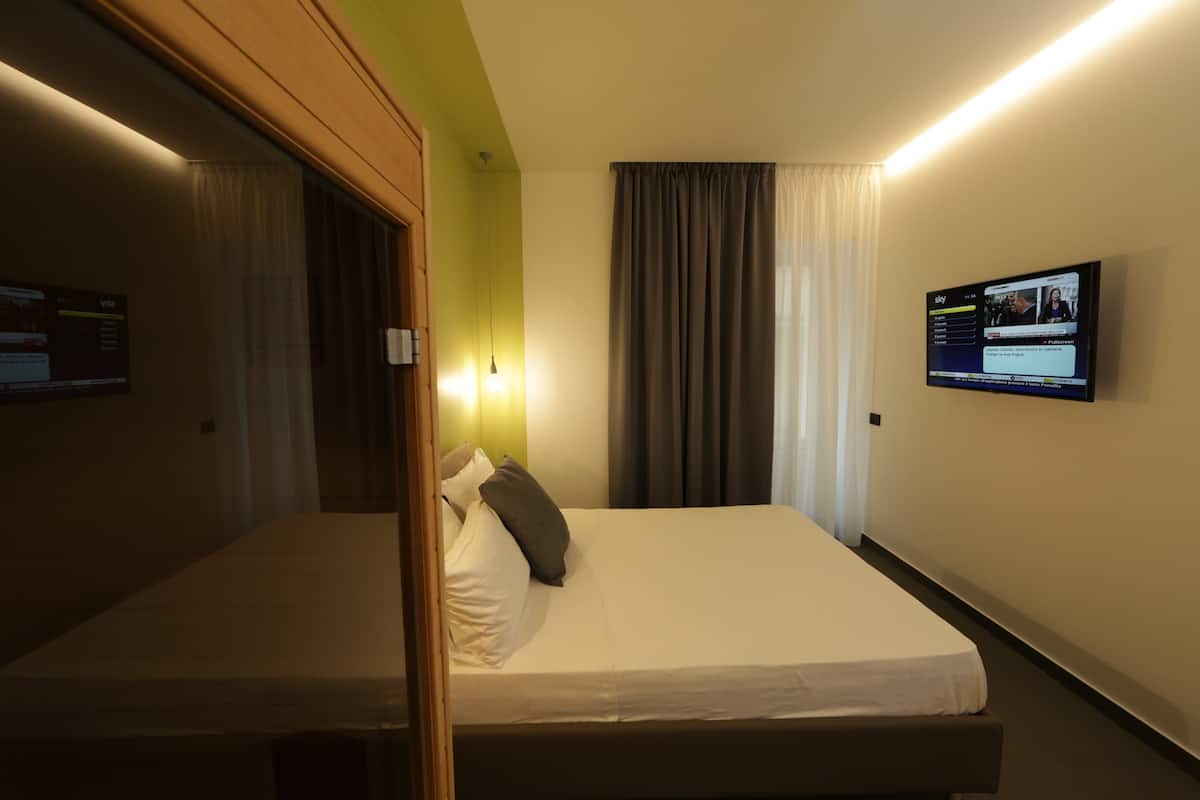 A hotel room with a single bed, a TV mounted on the wall, and a window with curtains.
