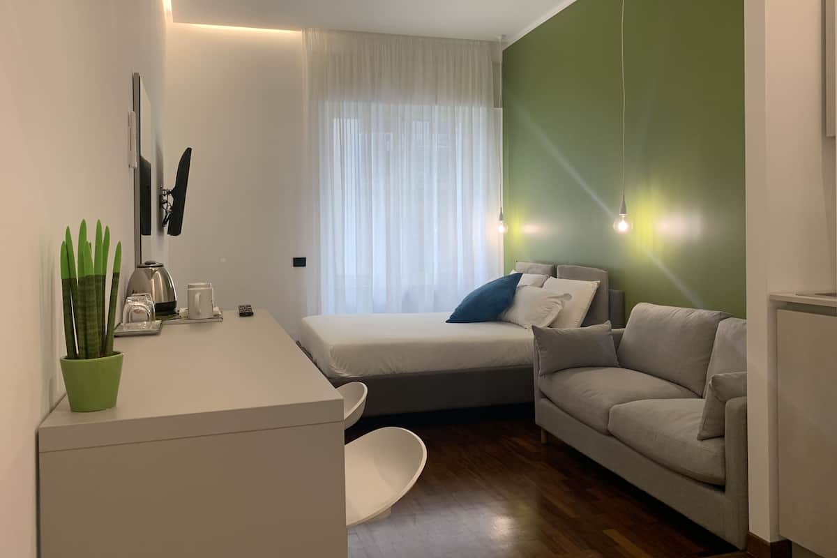 A modern bedroom with a bed, a sofa, a small table with a plant, and a window with sheer curtains.