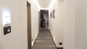 A narrow hallway with a light fixture on the left, a door on the left, and a wall with a textured artwork on the right. - Cellamare Suite & Spa (Naples)
