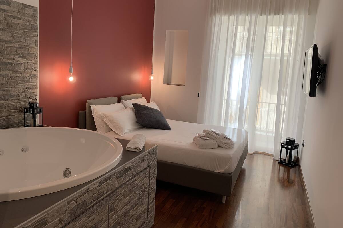 A bedroom with a large bathtub, a bed with white and gray bedding, and a stone-textured vanity.