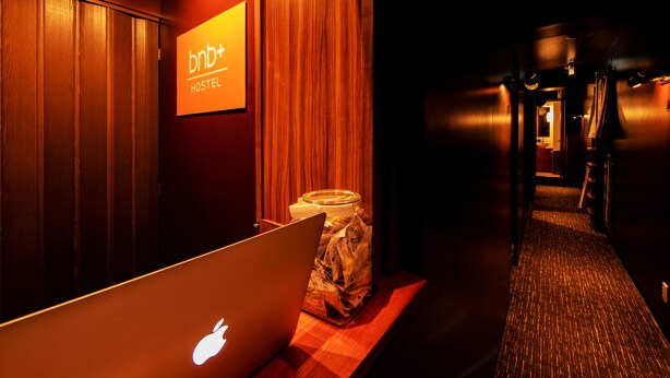 A hotel corridor with a wooden wall, a sign reading "bnb+ HOSTEL", and a laptop on a table. - bnb+ Ueno Okachimachi - Hostel (Tokyo)