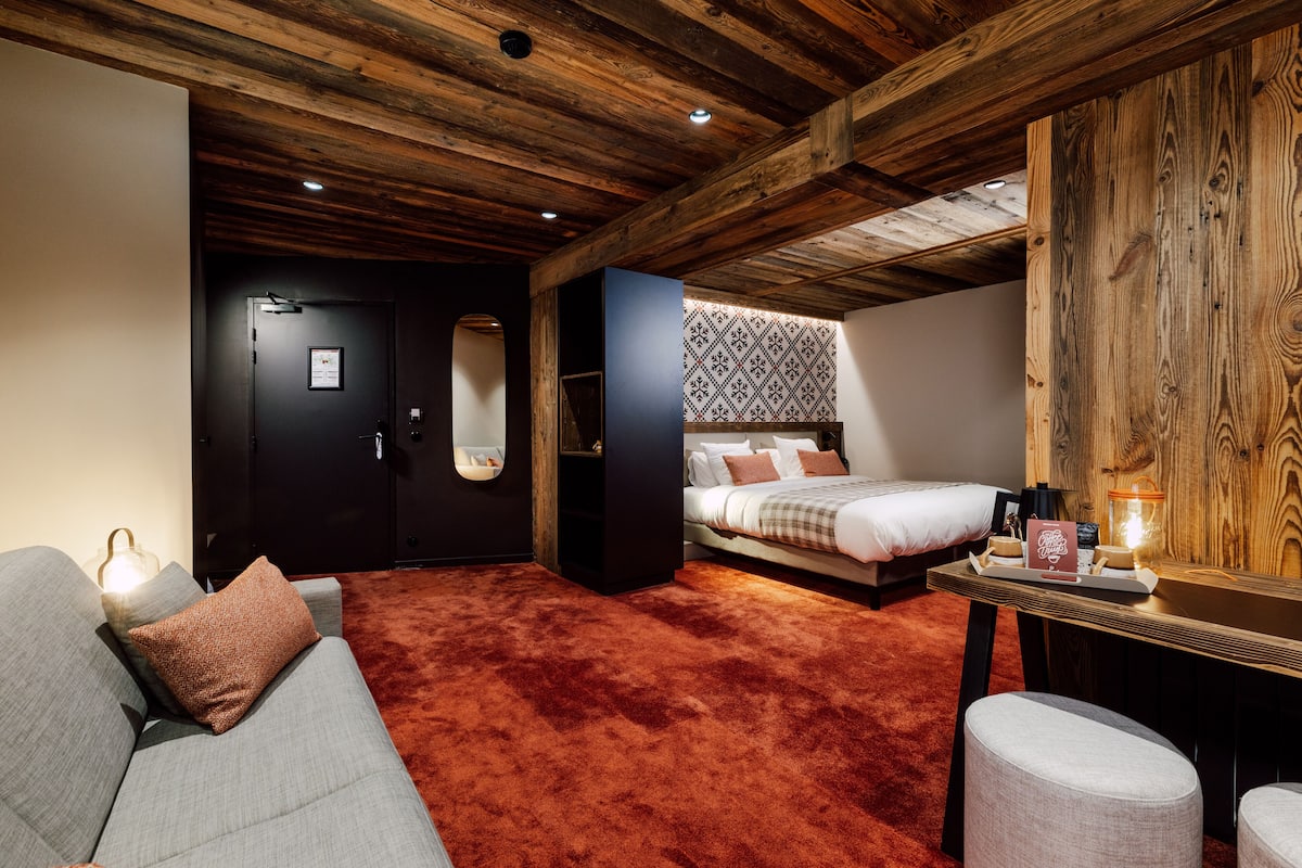A cozy bedroom with a wooden ceiling, a bed with patterned bedding, a grey sofa, and a bedside table with a lamp.