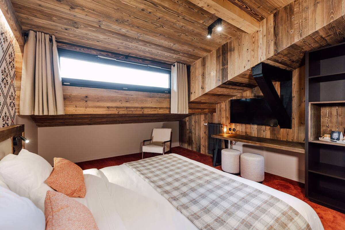 A bedroom with a wooden ceiling, a bed with white and orange pillows, a chair, a small table with a lamp, and a window with curtains.