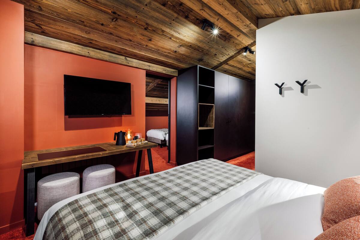A bedroom with a wooden ceiling, a TV mounted on the wall, a bed with a plaid bedspread, and a desk with a chair.