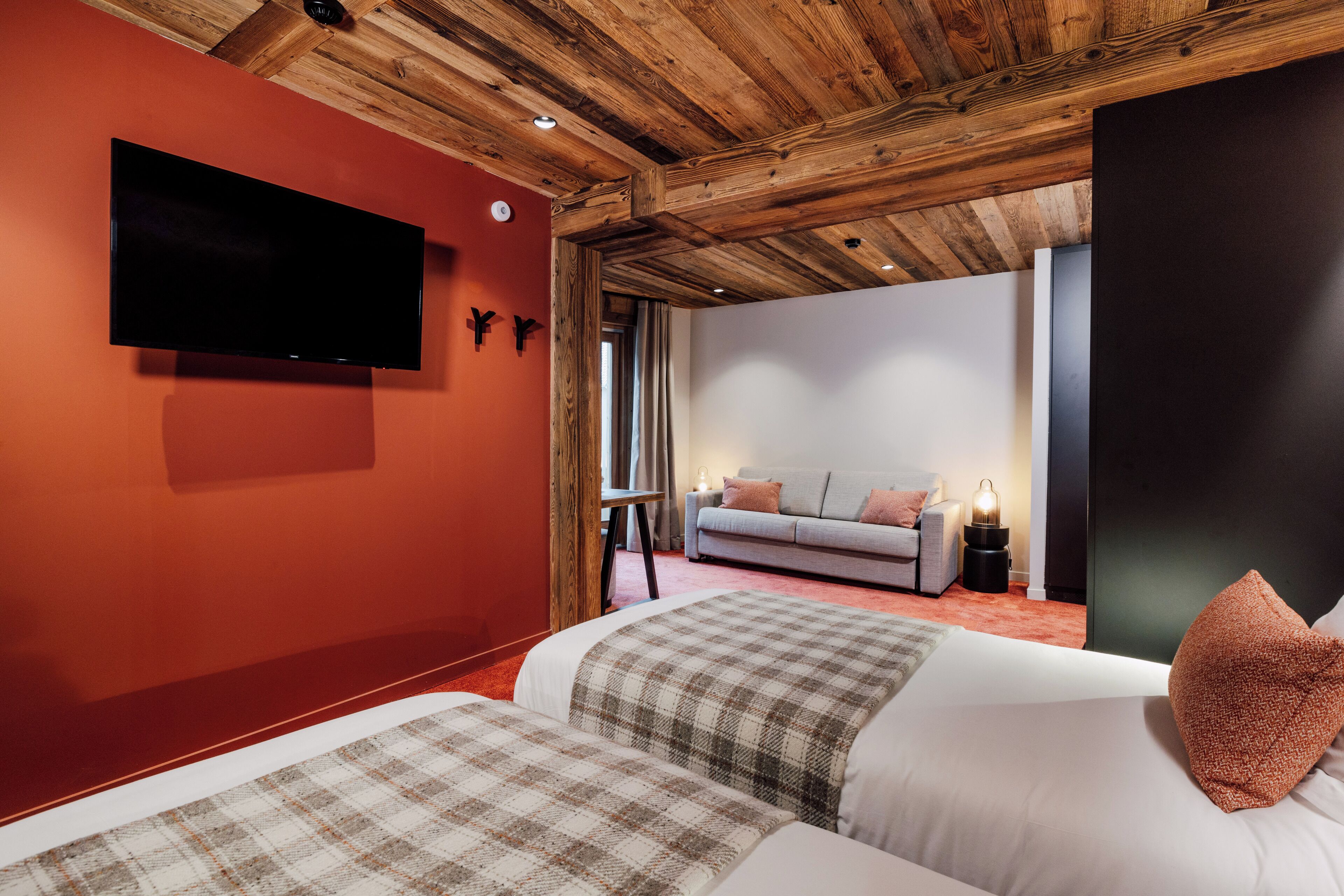 A hotel room with a wooden ceiling, a flat-screen TV mounted on the wall, a sofa, and a bed with a plaid blanket.