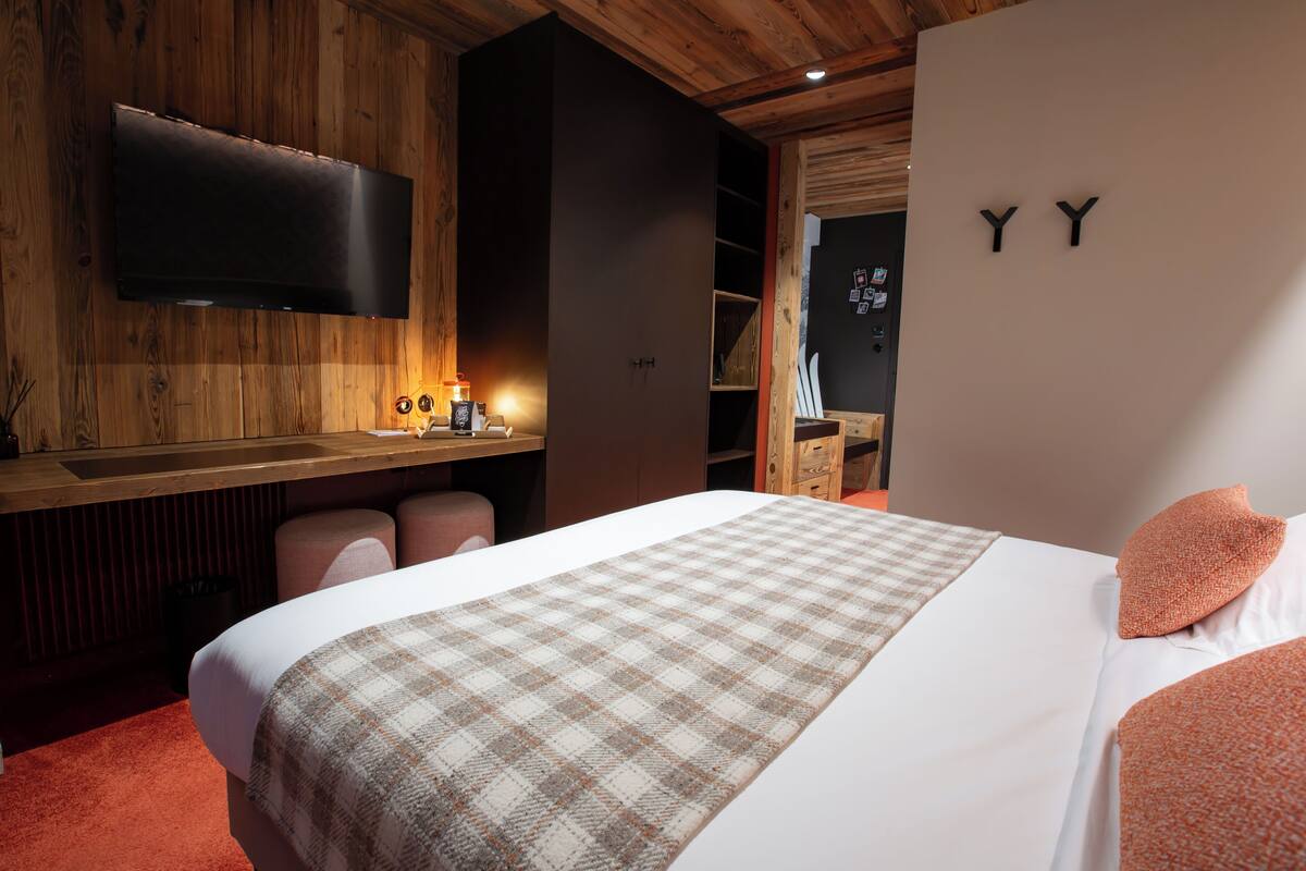 A modern hotel room with a large bed, a flat-screen TV mounted on the wall, and a wooden ceiling.