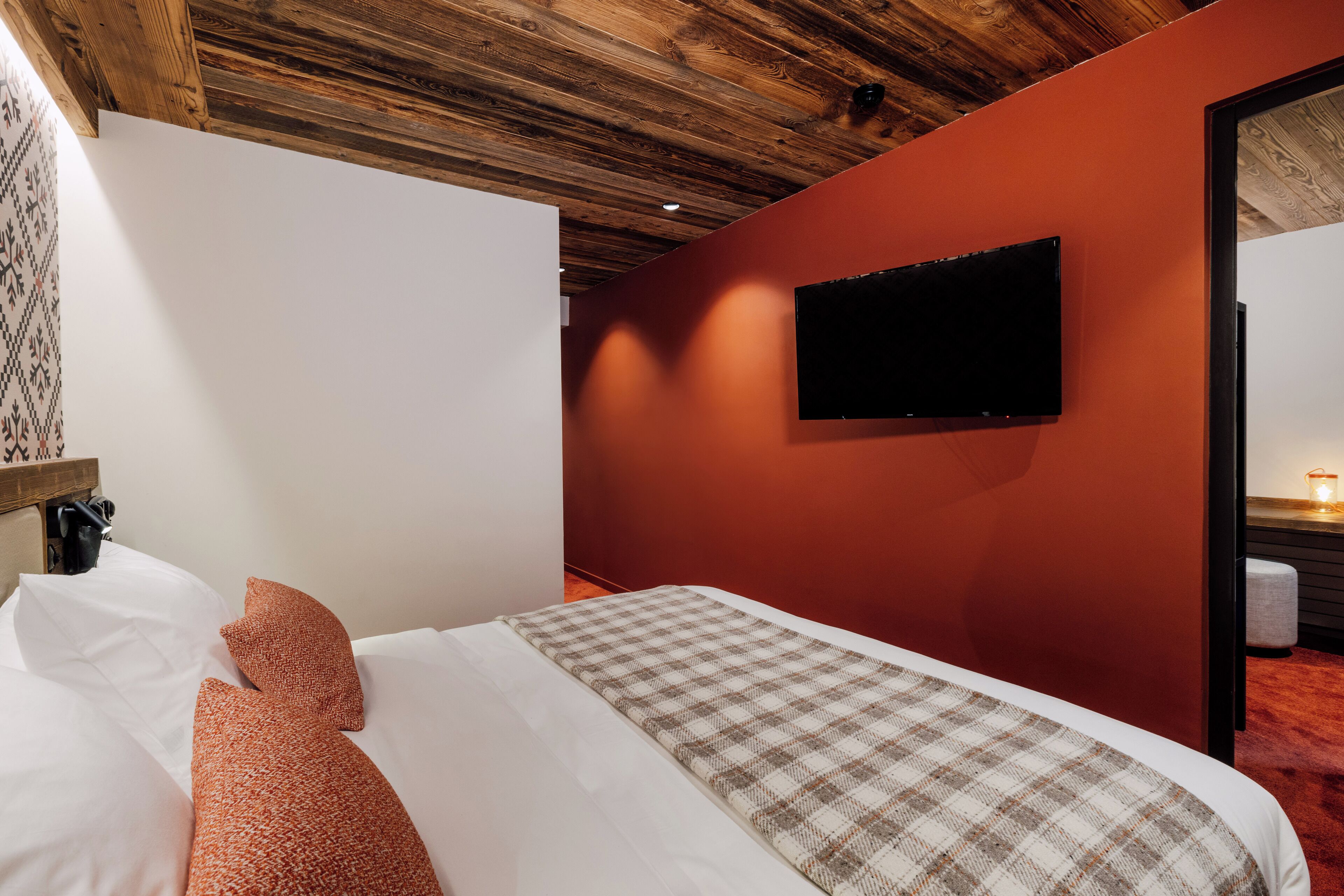 A hotel room with a wooden ceiling, a red accent wall, a flat-screen TV mounted on the wall, a bed with white and orange pillows, and a checkered throw blanket.