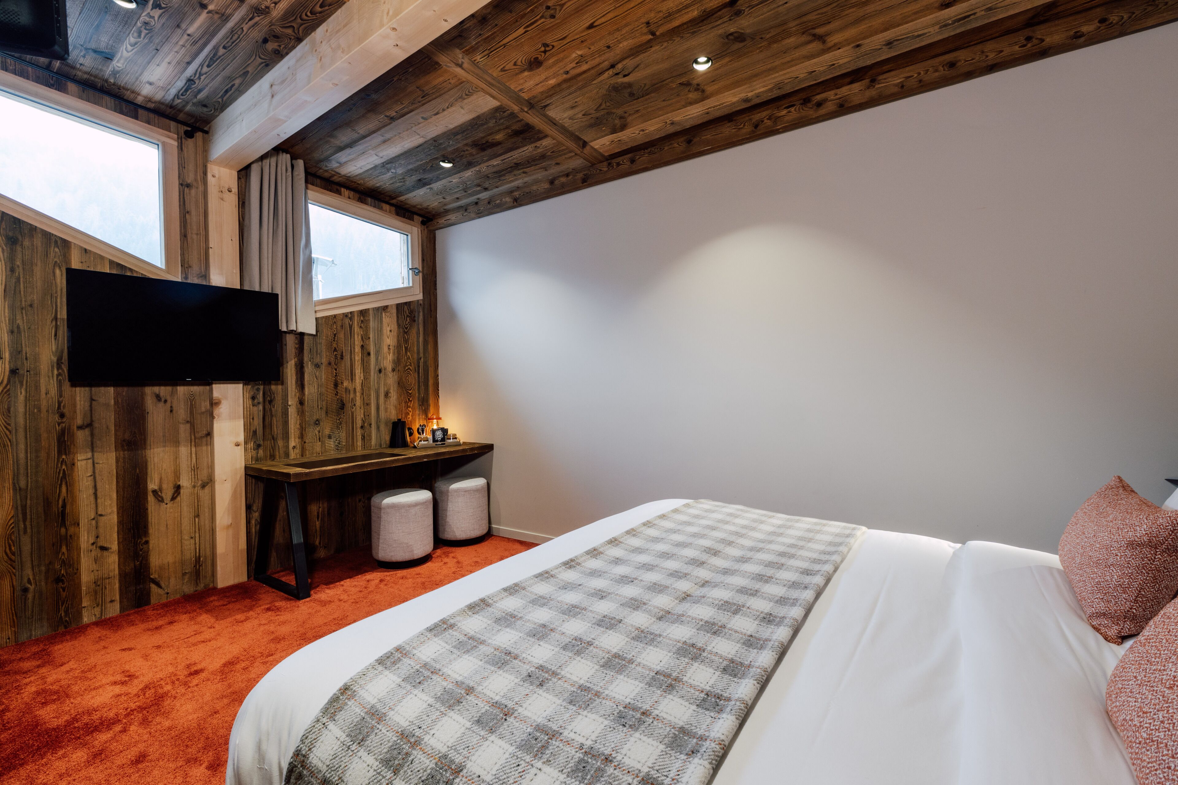 A bedroom with a wooden ceiling, a flat-screen TV mounted on the wall, a bed with a plaid blanket, and a desk with a lamp.