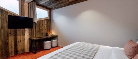 A bedroom with a wooden ceiling, a flat-screen TV mounted on the wall, a bed with a plaid blanket, and a desk with a lamp.