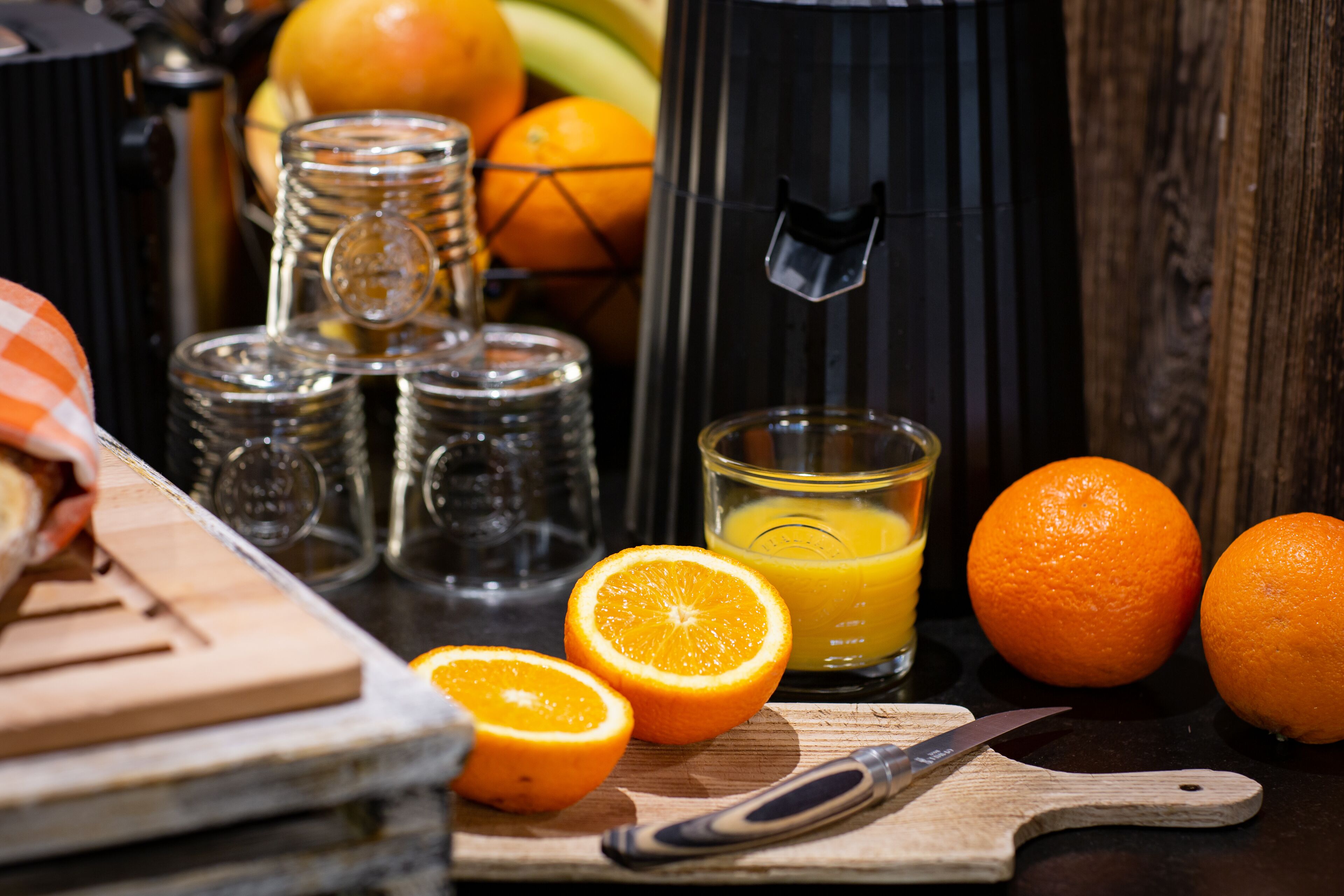 Eine Küchenarbeitsplatte mit Orangen, einem Glas Orangensaft, einem Messer und einer Kaffeemaschine.