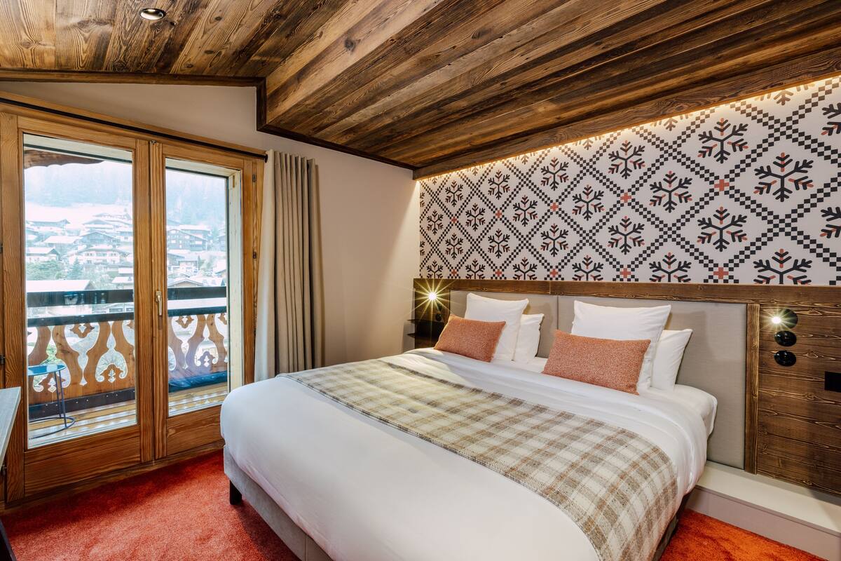 A hotel room with a large bed, wooden ceiling, patterned wallpaper, and a balcony with a view.
