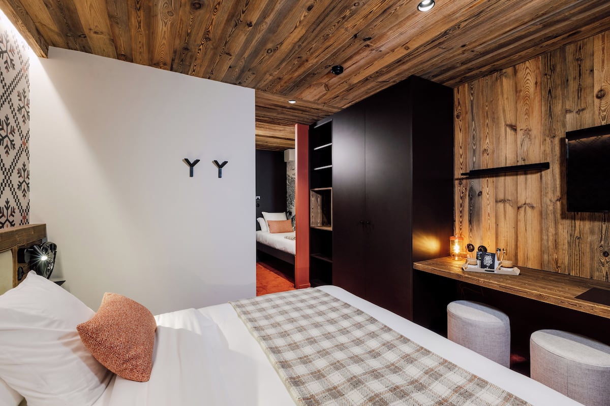 A modern bedroom with a wooden ceiling, a bed with white and brown bedding, a small desk with a lamp, and a wardrobe with a mirror.