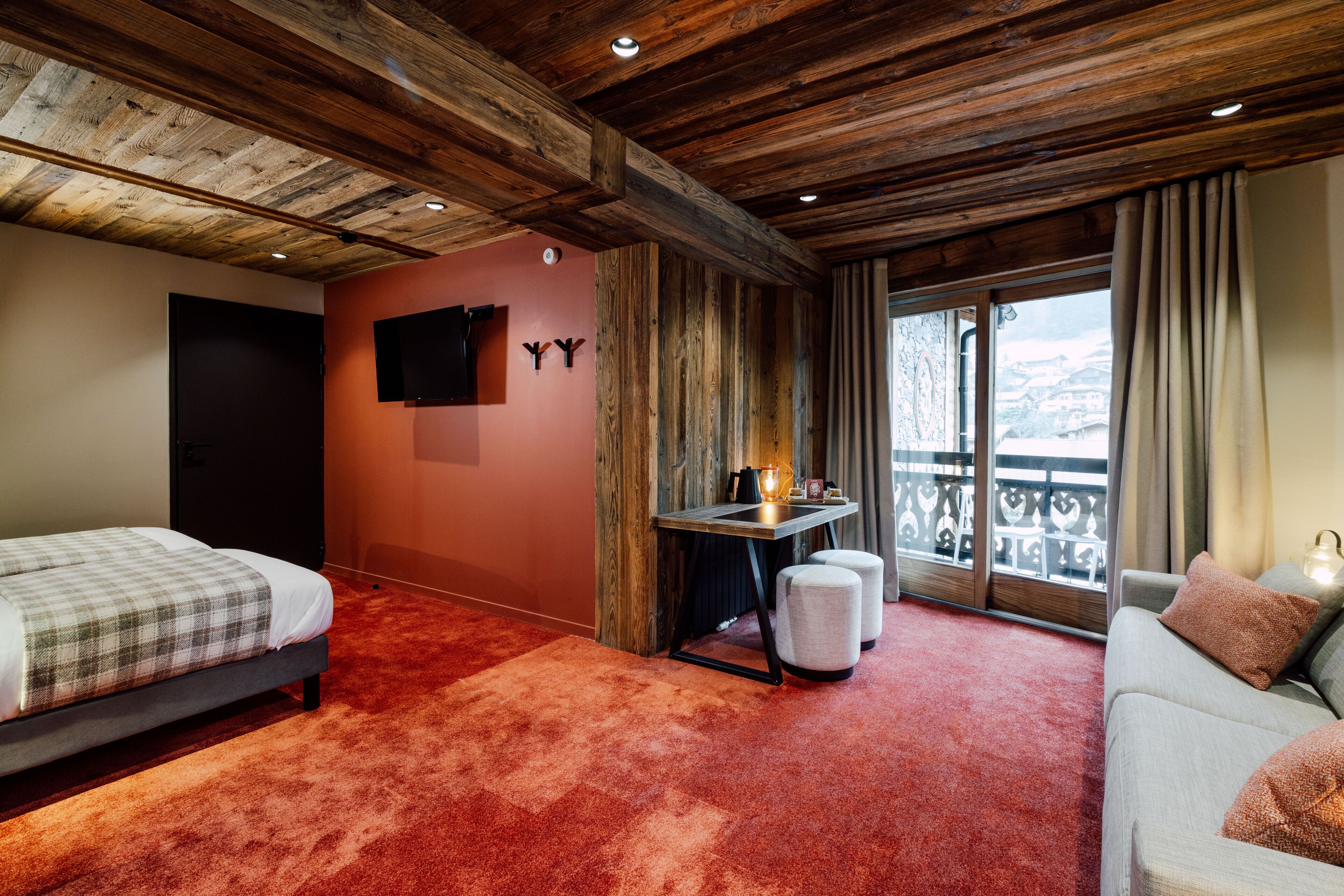 A bedroom with a wooden ceiling, a bed, a sofa, a small table with a lamp, and a window with a view of the outside.