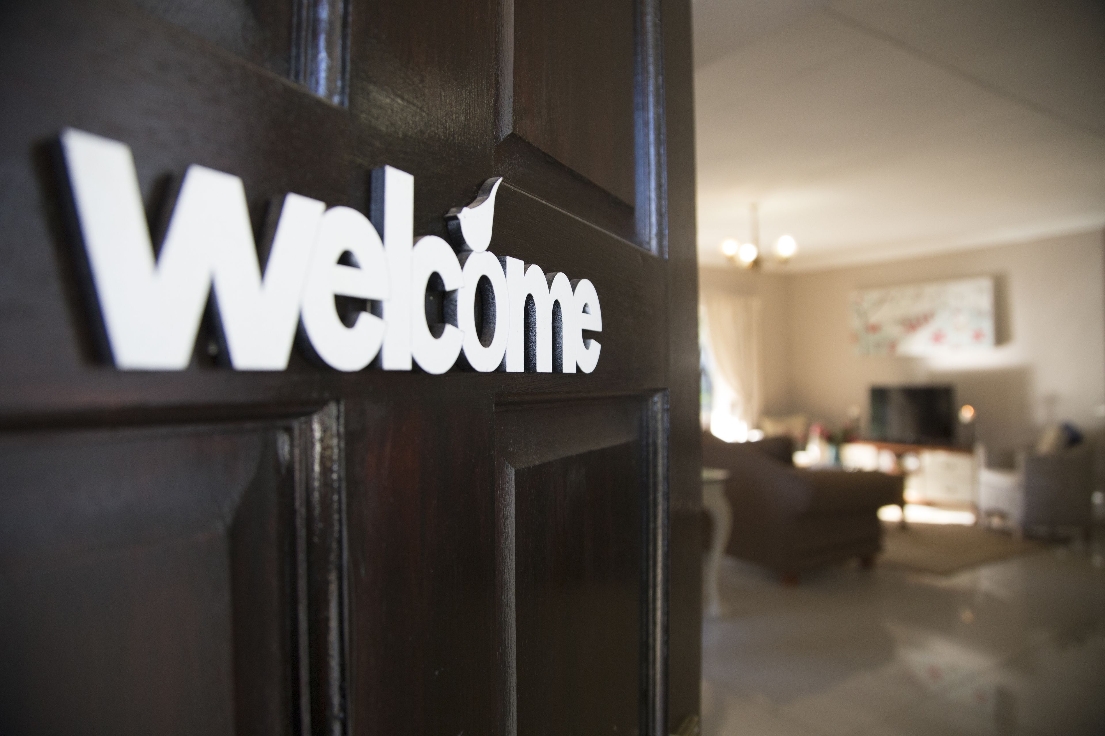 A 'Welcome' sign on a dark wooden door, with a blurred interior featuring a sofa and a television.