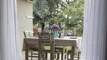 An outdoor dining area with a table set for four, surrounded by chairs, and a view of a garden with trees and a brick wall.
