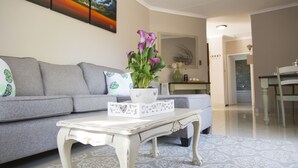 Ein Wohnzimmer mit einem grauen Sofa, einem weißen Couchtisch mit floralem Muster und einer Vase mit lila Blumen.