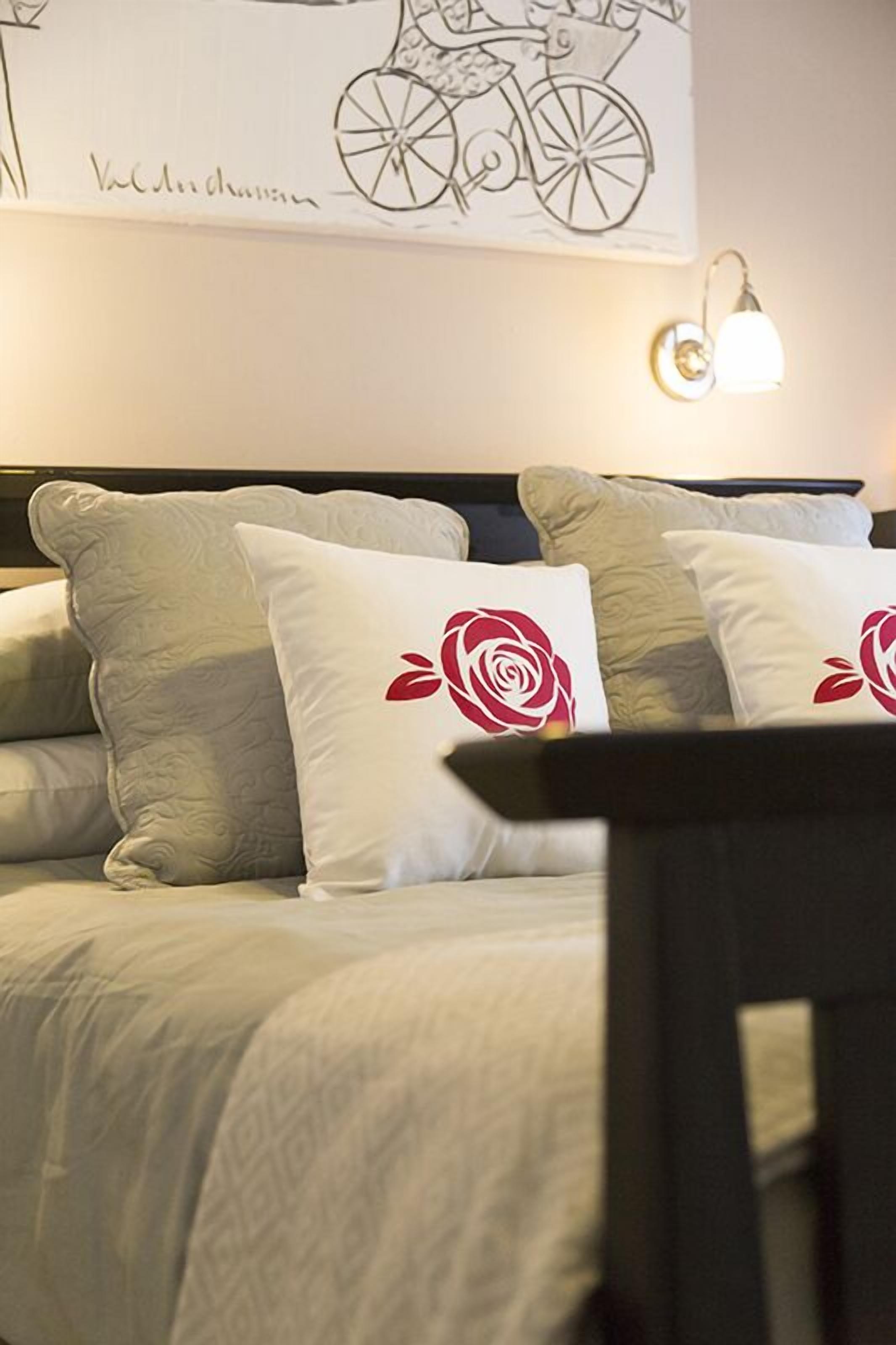 Un lit recouvert d’une couvre-lit à motifs, deux oreillers ornés de roses rouges et un luminaire fixé au mur.