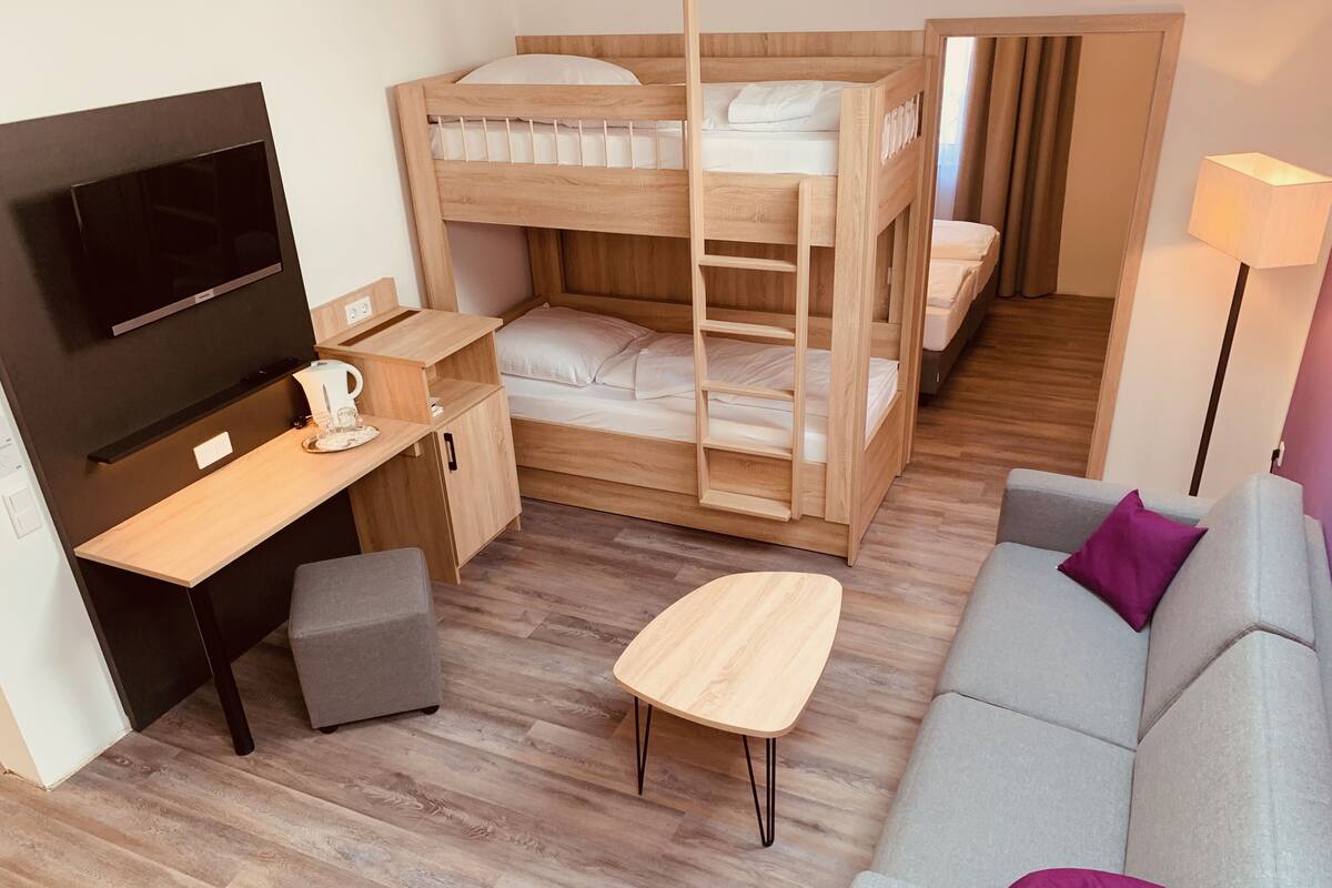 A compact room with a bunk bed, a desk with a TV, a grey sofa, and a small table.