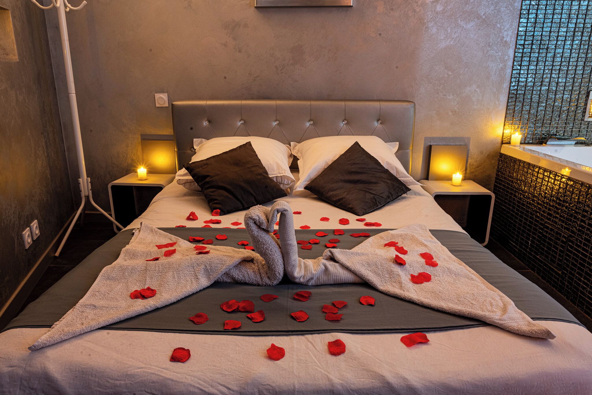 Una cama con pétalos de rosa y una toalla con forma de cisne, dos almohadas y lámparas de noche.