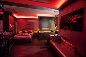A modern hotel room with a red-lit ambiance, featuring a bed, a sofa, a small table, and a wall-mounted TV. - Suite Romantique (Narbonne)