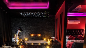 A dimly lit room with a stage, a bed, and a dining area with a table set for two. - Suite Romantique (Narbonne)