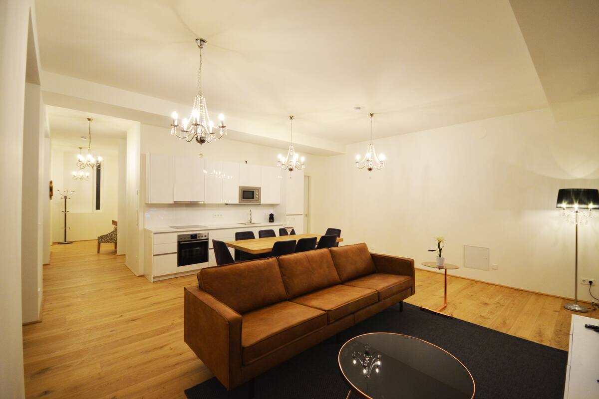 A modern living room with a brown sofa, a glass coffee table, and a dining area with a wooden table and chairs. There are two chandeliers hanging from the ceiling.