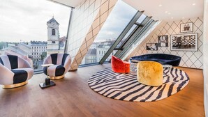 A modern living room with a panoramic city view, featuring a high ceiling, geometric wall design, and contemporary furniture. - Luxury Residence near Parliament (Vienna)