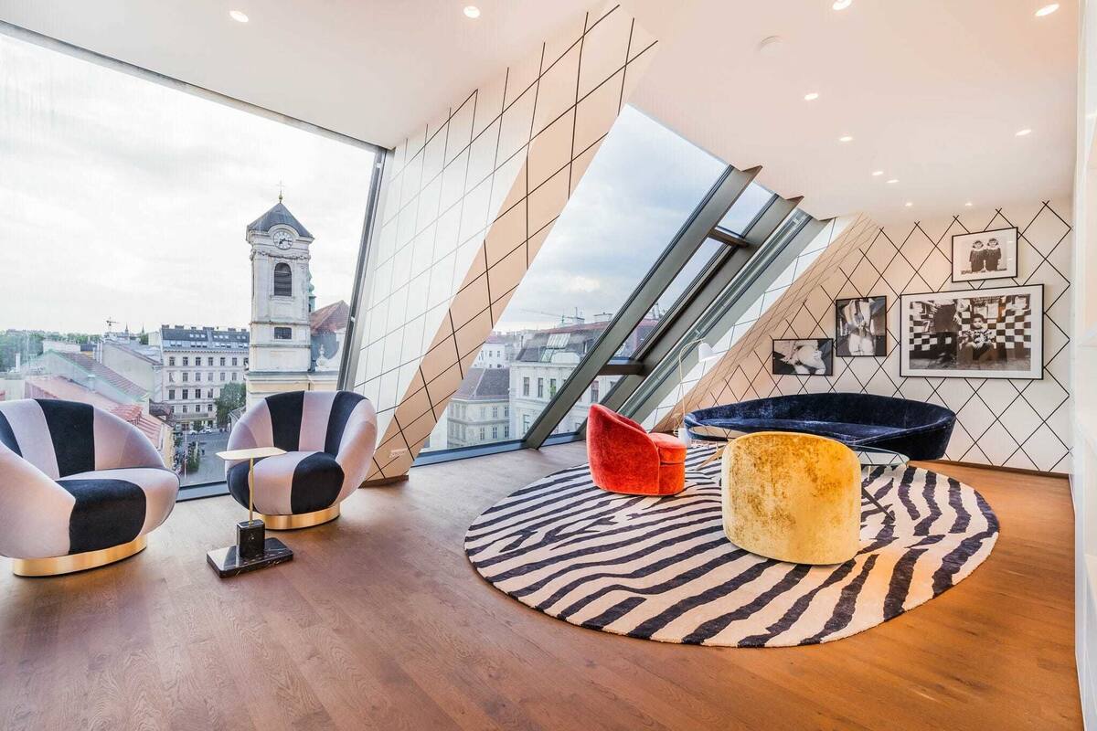 A modern living room with a panoramic city view, featuring a high ceiling, geometric wall design, and contemporary furniture.