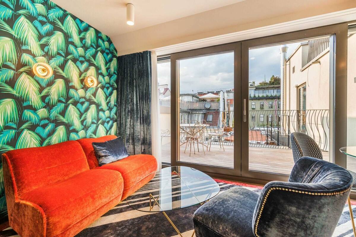A modern living room with a vibrant green and blue leaf-patterned wall, a red sofa, a glass coffee table, and a view of the city through large windows.