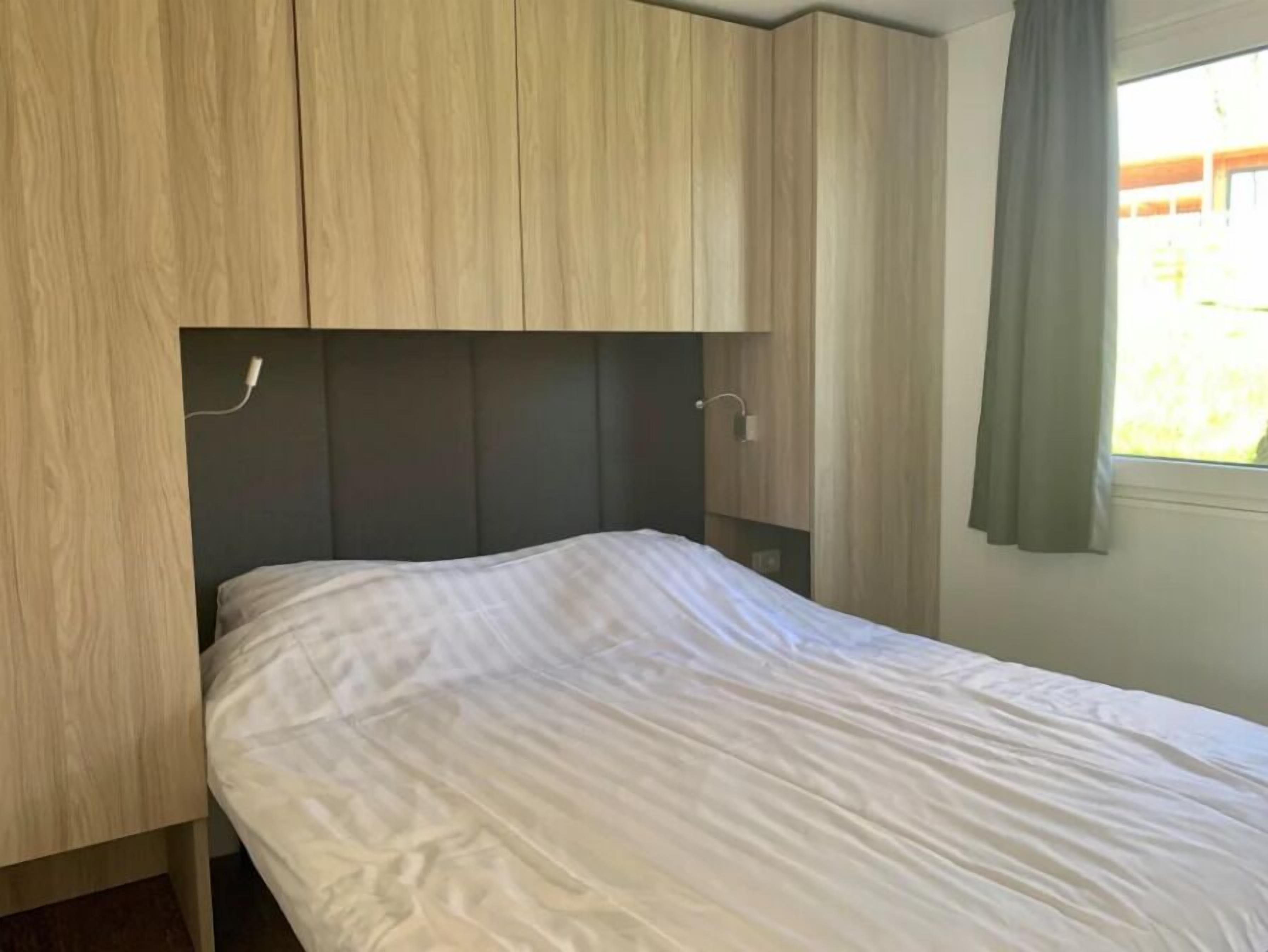 A single bed with a headboard against a wall, a window with curtains, and wooden paneling on the wall.