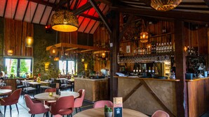 Un interior rústico con vigas de madera, mesas y sillas redondas. Se observa una zona de bar con estantes llenos de botellas y vasos.