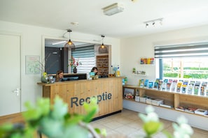 A reception area with a wooden desk labeled "Receptie," a woman behind the desk, and a window with bookshelves. - Europarcs Gulperberg (Gulpen)