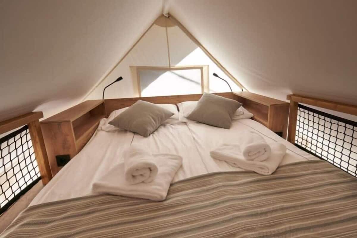 Una cama individual con cabecera y piecera de madera, una ventana en la parte superior y una barandilla con mosquitero a los lados.