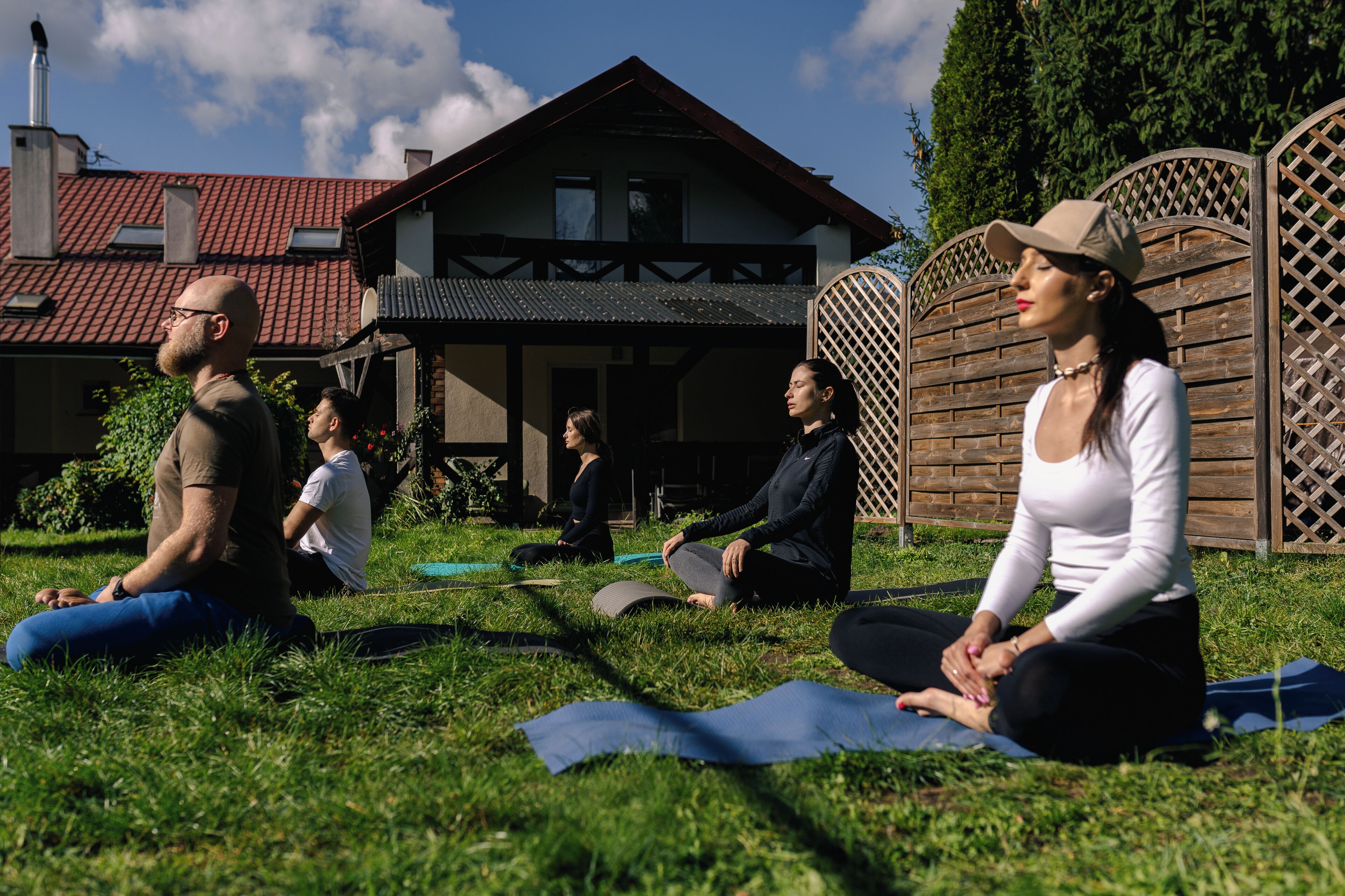A group of people sitting on yoga mats outdoors in a grassy area with a house in the background.