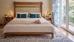 A bed with a wooden headboard, two bedside tables with lamps, a large window with a view of greenery, and a patterned rug. - Mignon & Teak (Athens)