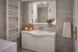 A bathroom with a white vanity, a mirror, a towel rack, and a towel hanging on the wall. - Mignon & Teak (Athens)