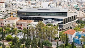 A modern building with a glass facade, surrounded by trees and older buildings. - Mignon & Teak (Athens)