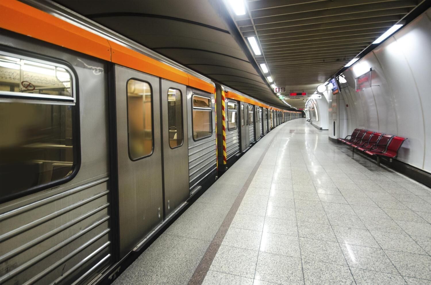 A subway station with a train on the left, tiled floor, and red benches on the right.