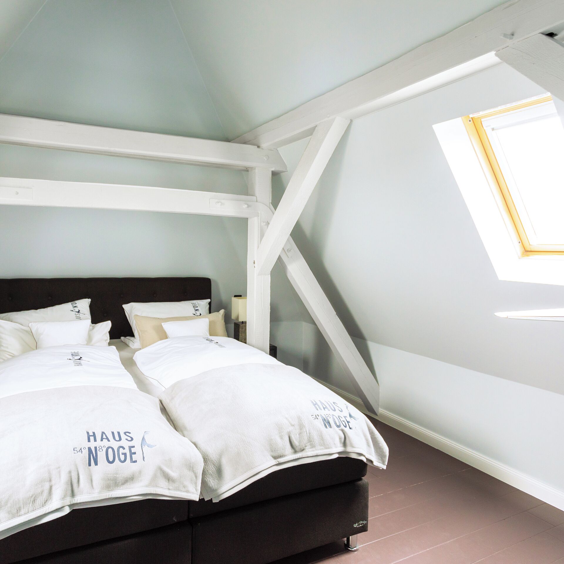Une chambre à coucher avec un lit, des oreillers, une table de chevet, une lampe et une lucarne.