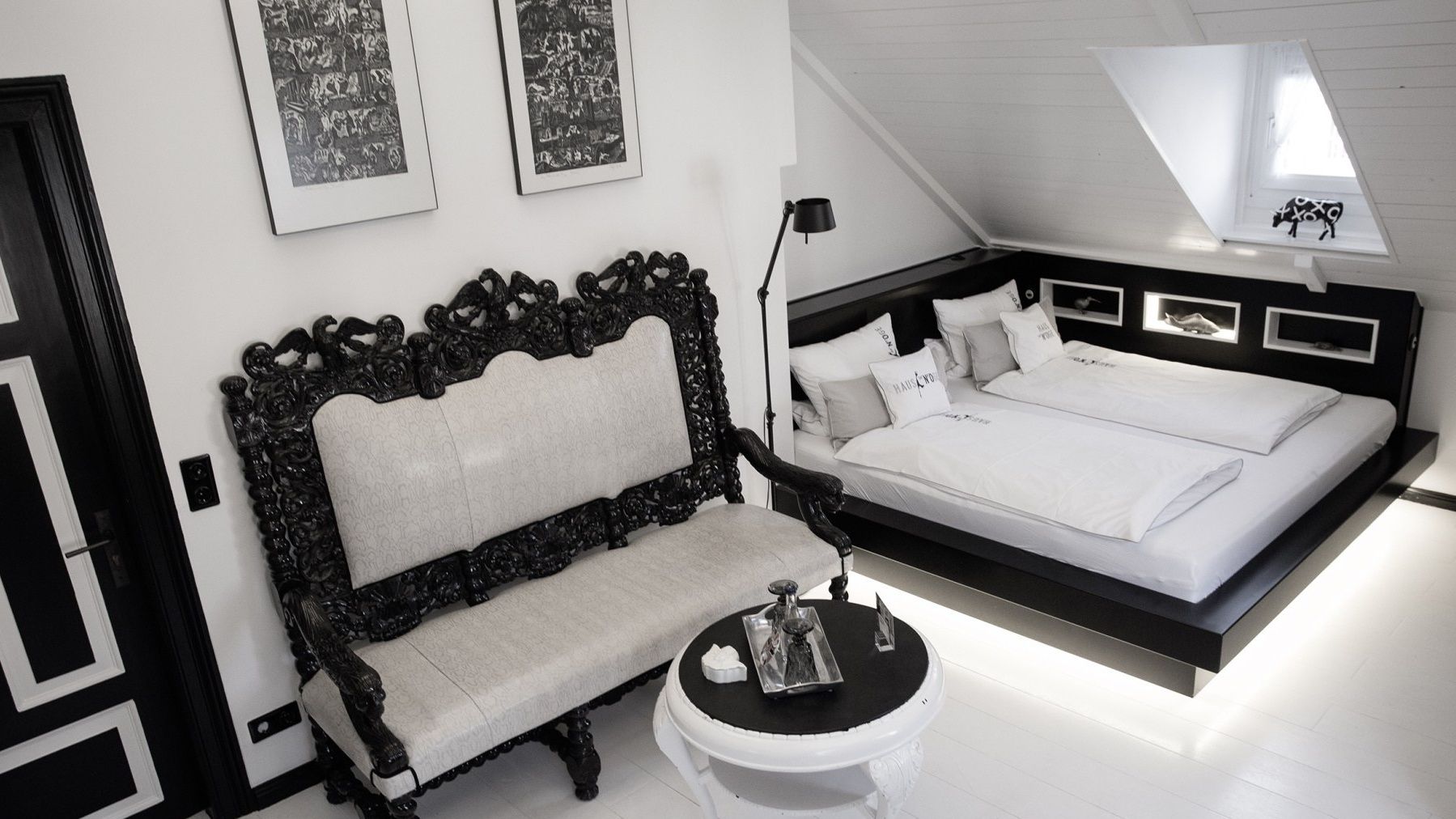 A modern bedroom with a large bed, a classic armchair, a small table, and two framed pictures on the wall.
