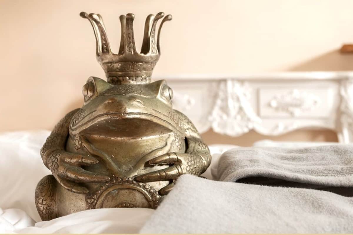 A decorative frog statue with a crown on a bed with a white headboard.