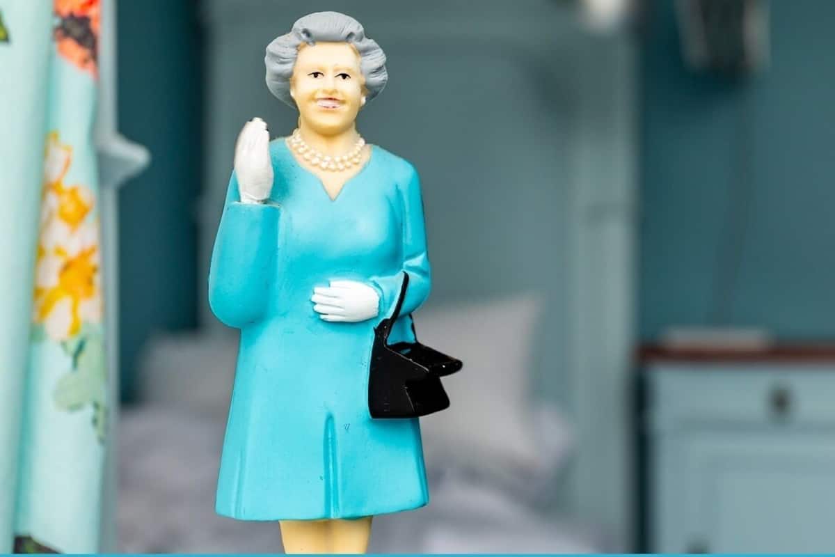 A figurine of a woman in a blue dress and pearl necklace, standing in a room with a bed and a nightstand.