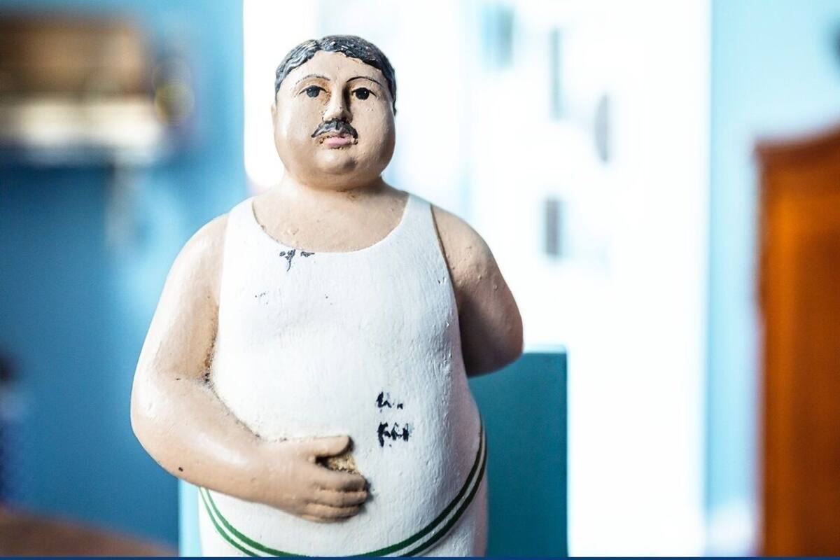 A statue of a person with a white tank top and green trim, standing in a room with a blue wall and a wooden door.