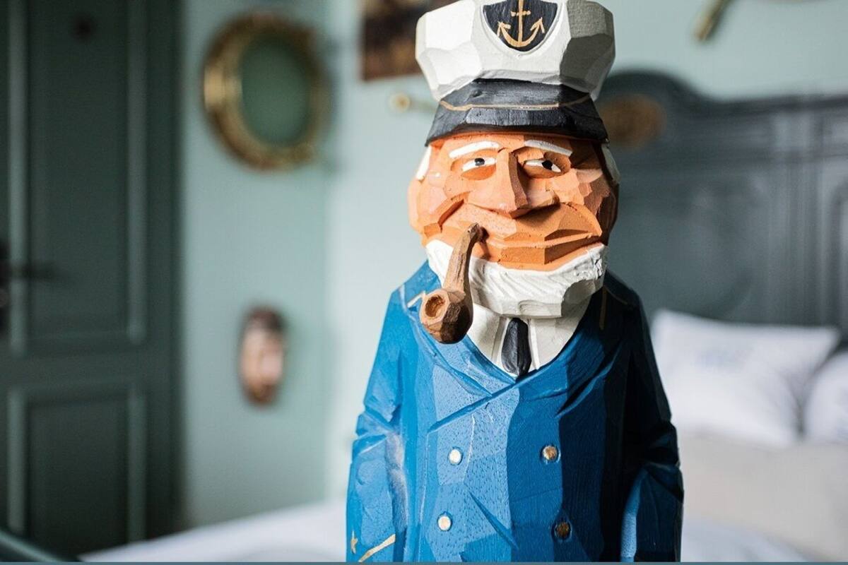 A wooden figure dressed as a sailor in a room with a bed, a door, and a wall-mounted circular object.
