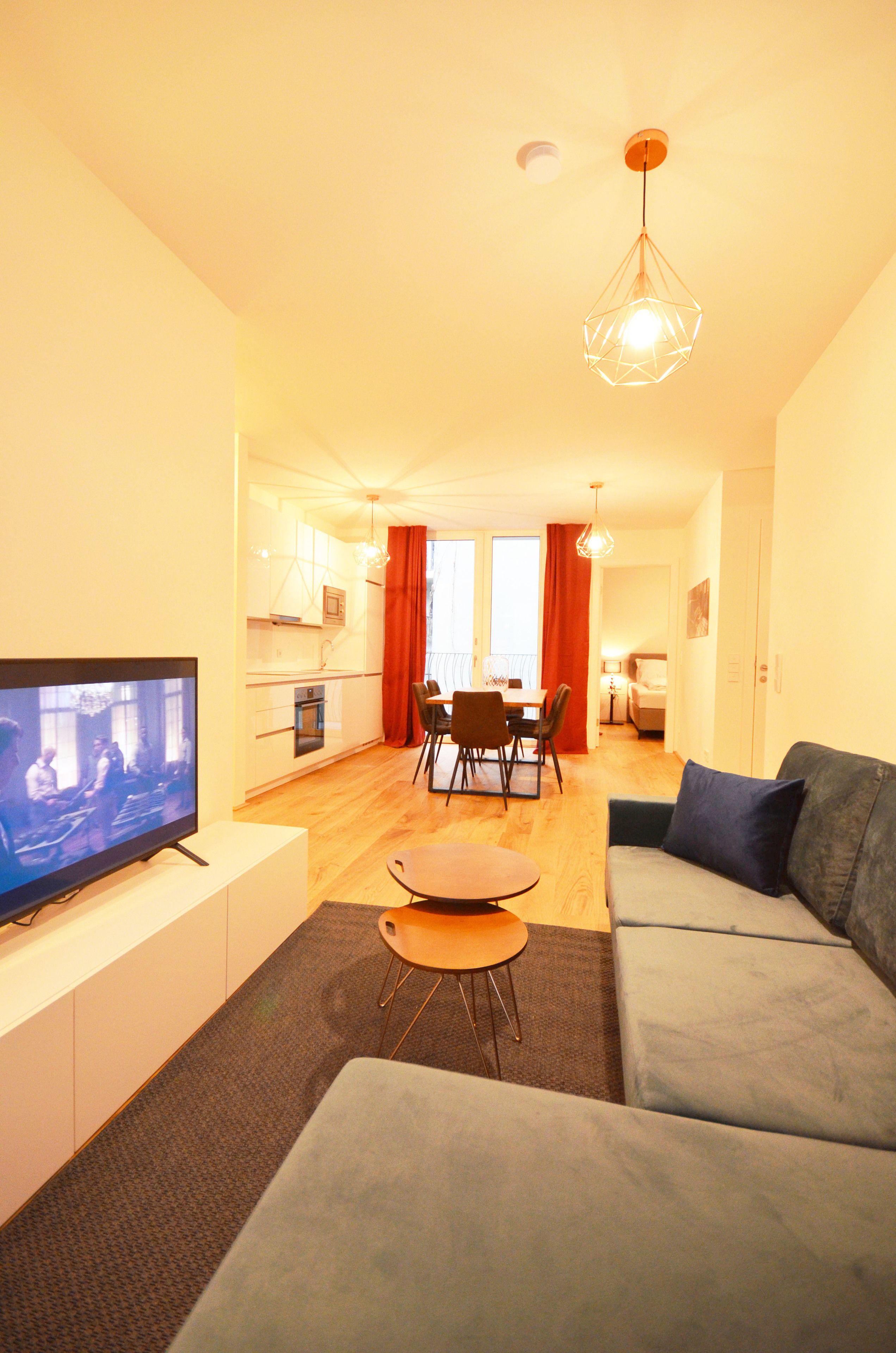 A modern living room with a flat-screen TV, a grey sofa, and a dining area with a table and chairs.